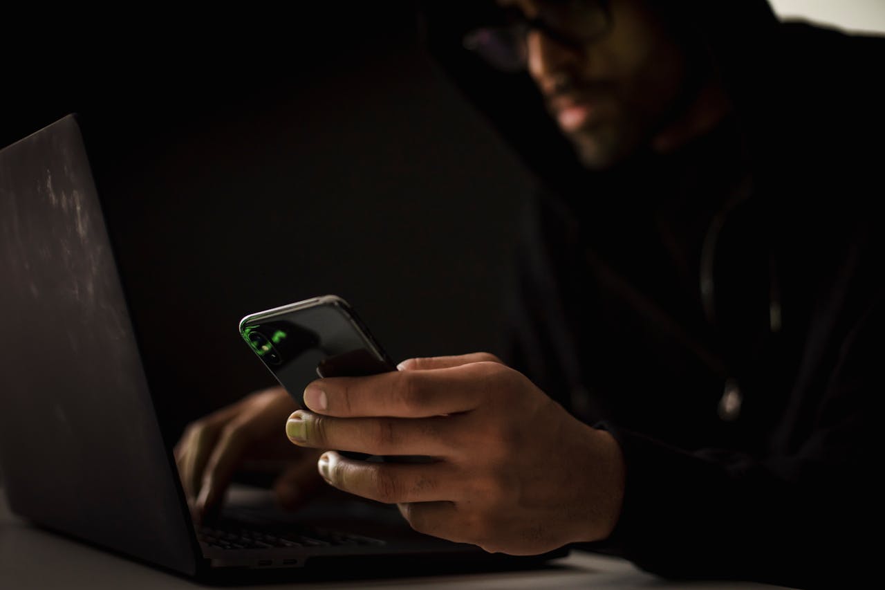 Crop anonymous ethnic male cyber spy with cellphone and netbook hacking system in evening